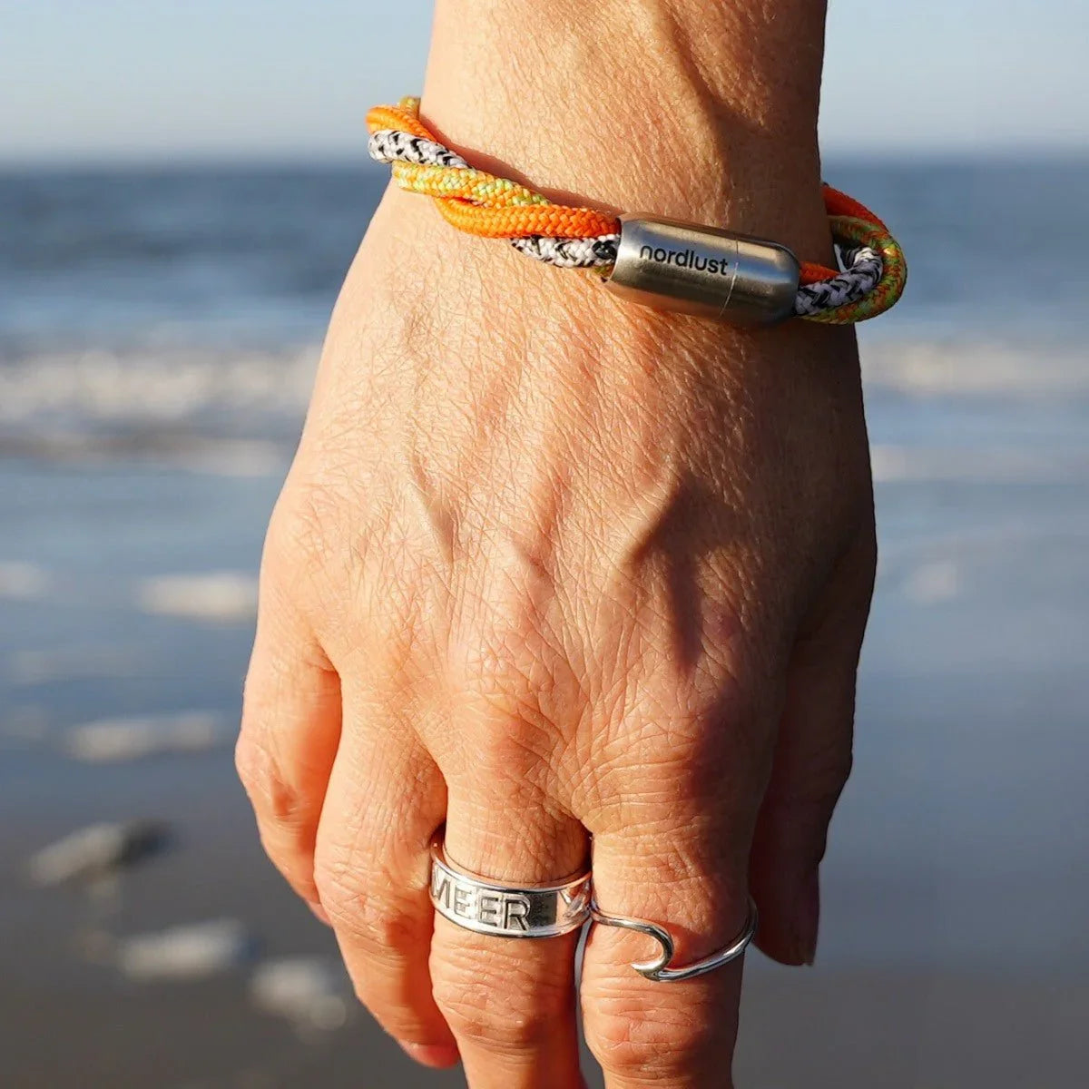 Maritimes Armband aus orange-gelbem Tau auf Hand am Strand mit Meer im Hintergrund