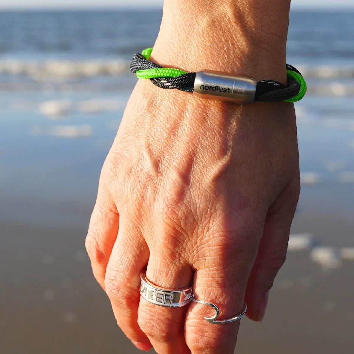 Hand mit maritimem Armband aus schwarzem und grünem Seil am Strand, Meer im Hintergrund
