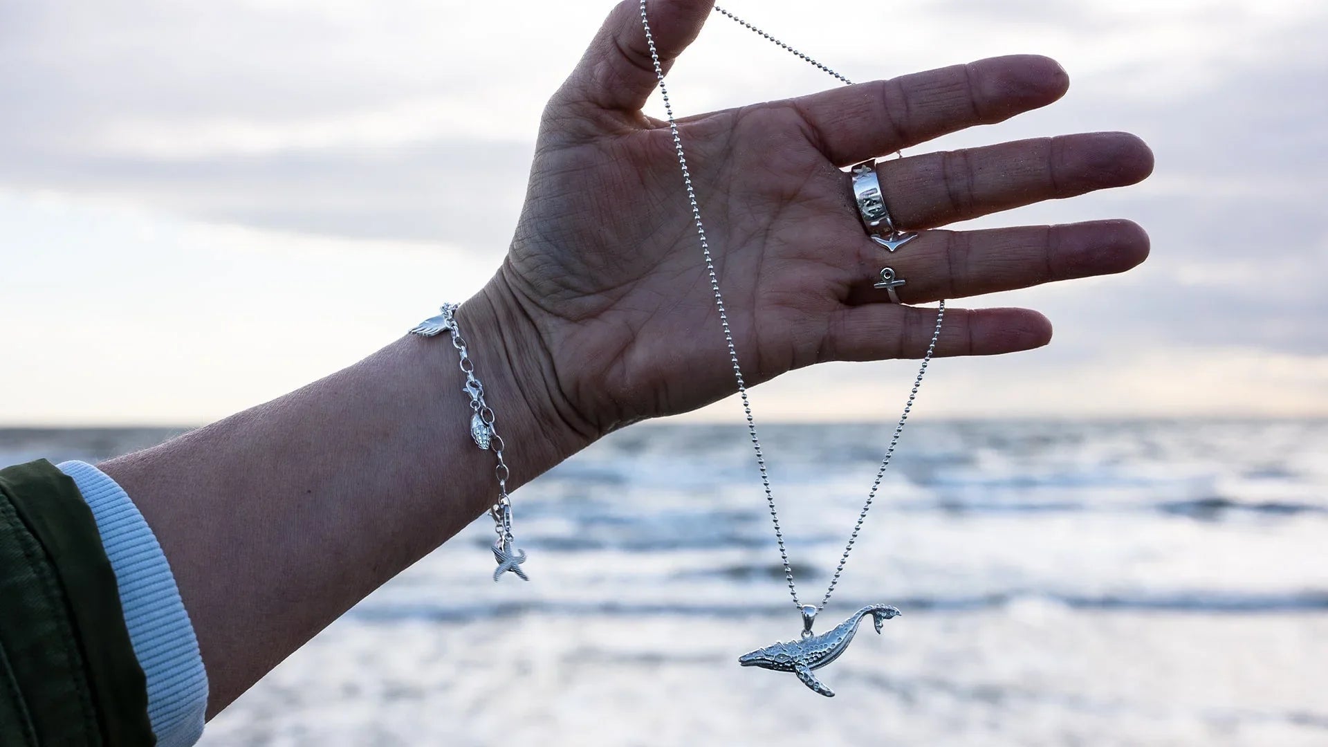 Silberkette mit Wal-Anhänger und maritimes Armband am Handgelenk vor dem Meer