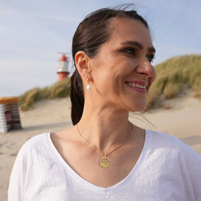 Frau am Strand mit goldener Ankerkette und Perlenohrring, maritimer Schmuck, Leuchtturm im Hintergrund