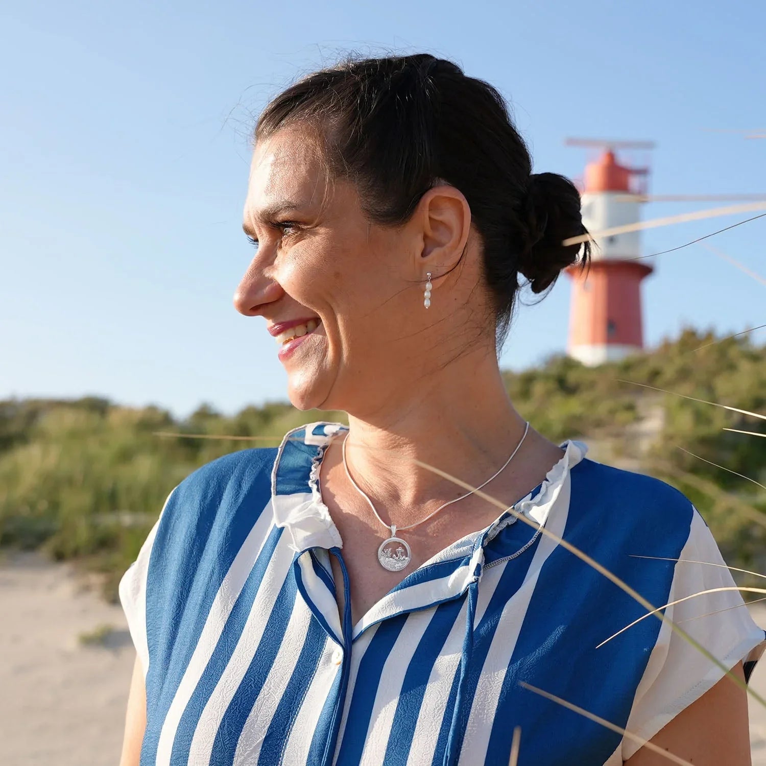 Frau mit maritimer Silberkette mit Anker-Anhänger am Strand, Leuchtturm im Hintergrund