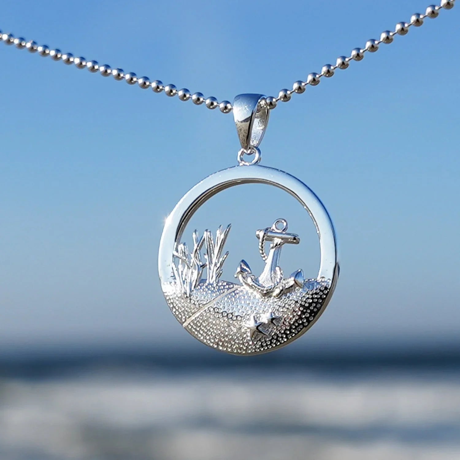 Silberner runder Anhänger mit Anker-Motiv, maritimer Schmuck am Strand vor blauem Himmel