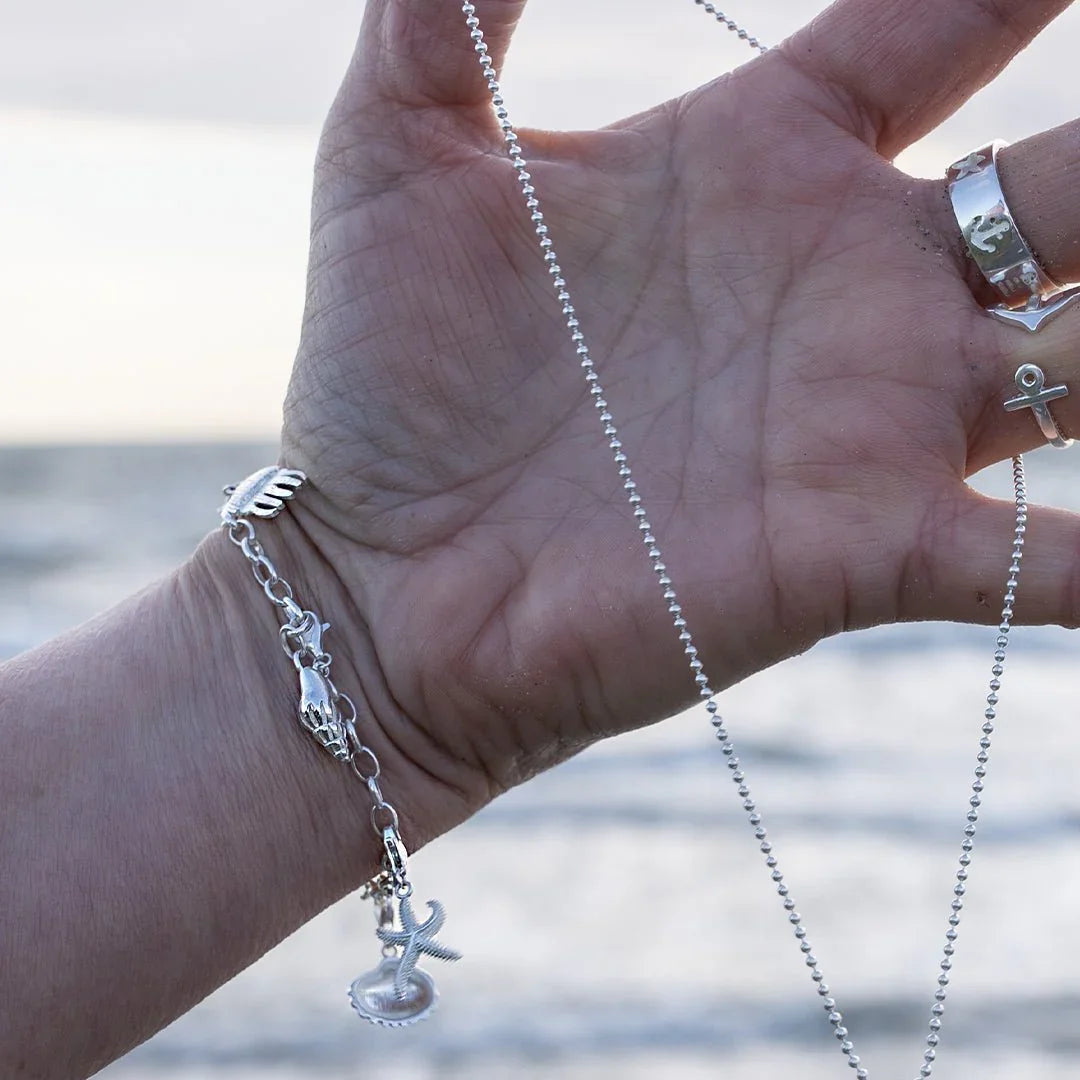 Silbernes Armband mit maritimen Anhängern wie Anker und Muschel am Handgelenk vor dem Meer