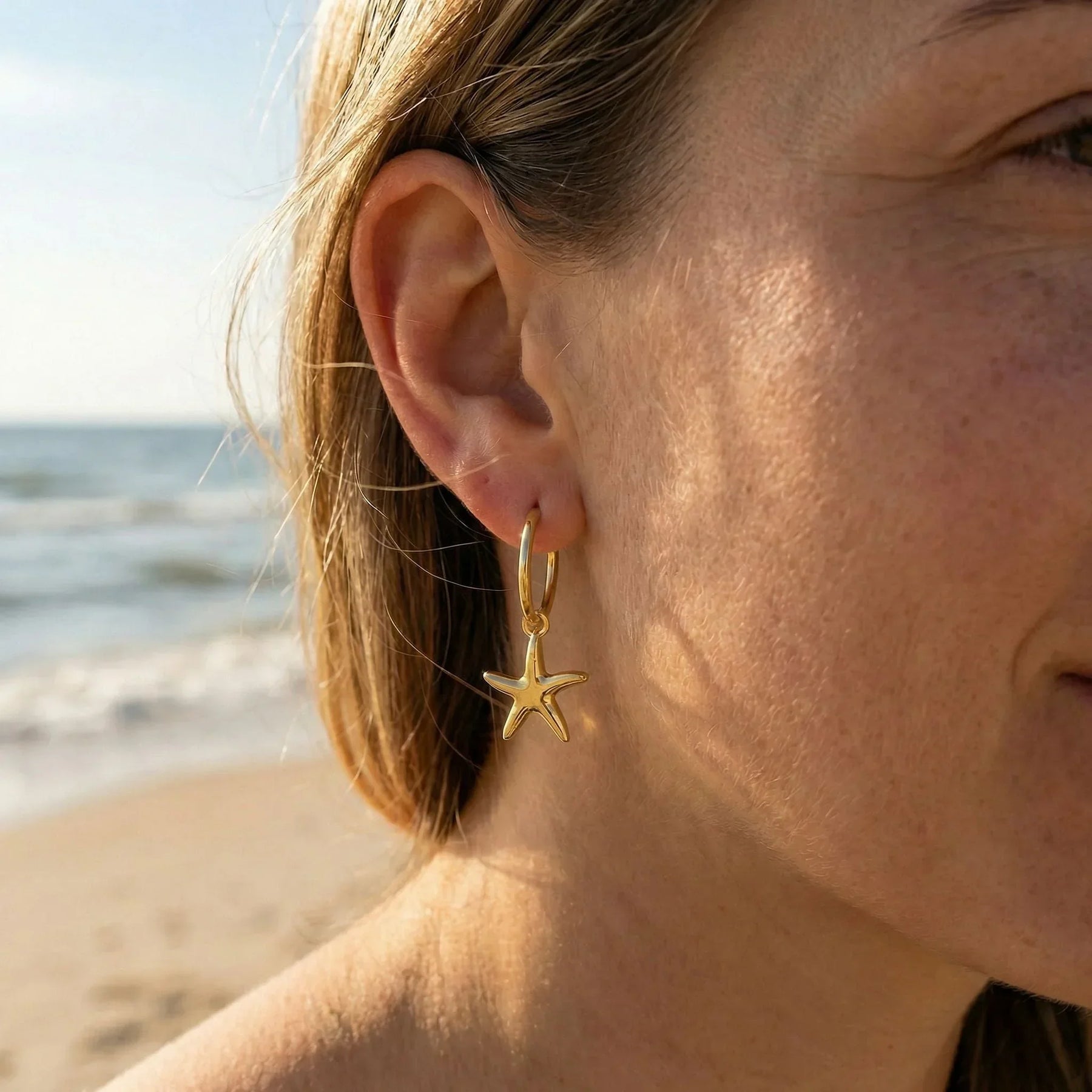 Goldene Creole mit Seestern-Anhänger am Ohr einer Frau, Strand und Meer im Hintergrund