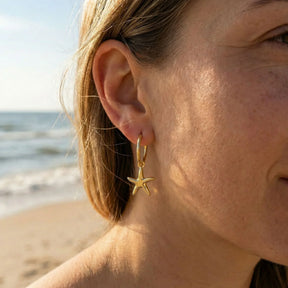 Goldene Creole mit Seestern-Anhänger am Ohr einer Frau, Strand und Meer im Hintergrund