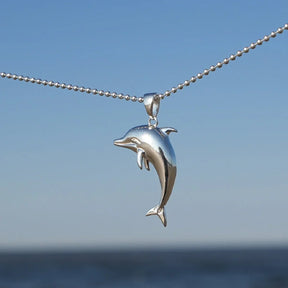 Silberner Delfin-Anhänger an Kette vor blauem Himmel und Meer, maritimer Schmuck