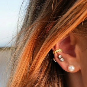 Ohr mit goldfarbenem Muschel-Earcuff, Perlenohrring und funkelnden Ohrsteckern am Ohr.