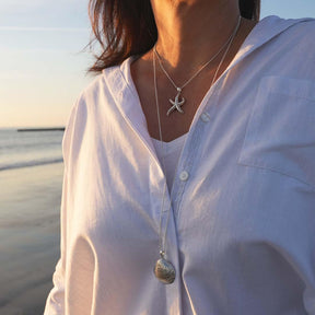 Frau am Strand mit weißer Bluse, trägt silberne Muschel- und Seestern-Anhänger-Ketten