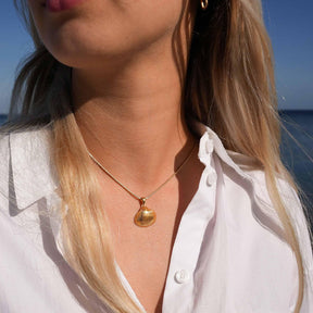 Frau am Strand mit goldener Herzmuschel-Kette und weißer Bluse, maritimer Schmuck