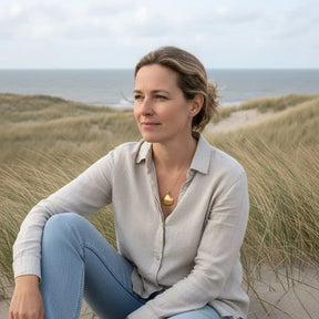 Frau mit goldener Herzmuschel-Kette sitzt in den Dünen am Meer