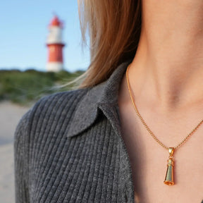 Goldene Leuchtturm-Kette am Hals, im Hintergrund Küste und roter Leuchtturm