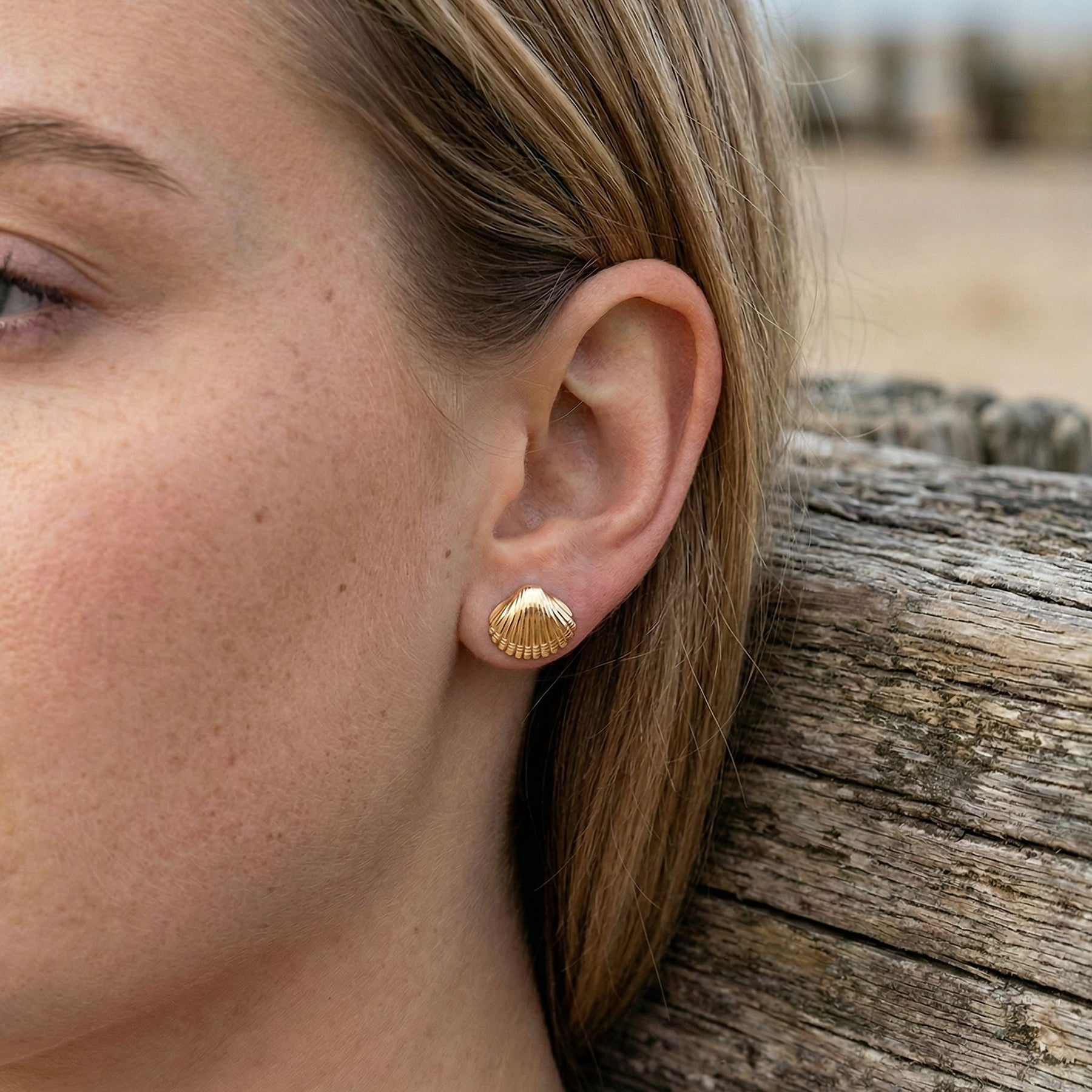Goldener Ohrstecker in Herzmuschel-Form am Ohr einer Frau am Strand