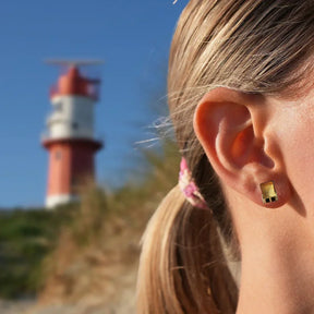 Goldene Ohrstecker in Strandkorb-Form am Ohr, maritimer Hintergrund mit Leuchtturm
