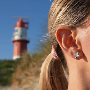 Ohrstecker aus Silber in Strandkorb-Form am Ohr, maritimer Schmuck, Leuchtturm im Hintergrund