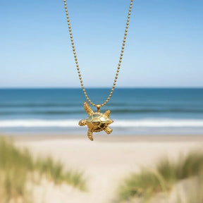 Goldene Schildkröte Anhänger Kette am Strand mit Meer im Hintergrund, maritimer Schmuck