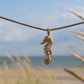 Goldener Seepferdchen-Anhänger an Kette vor maritimem Strand-Hintergrund