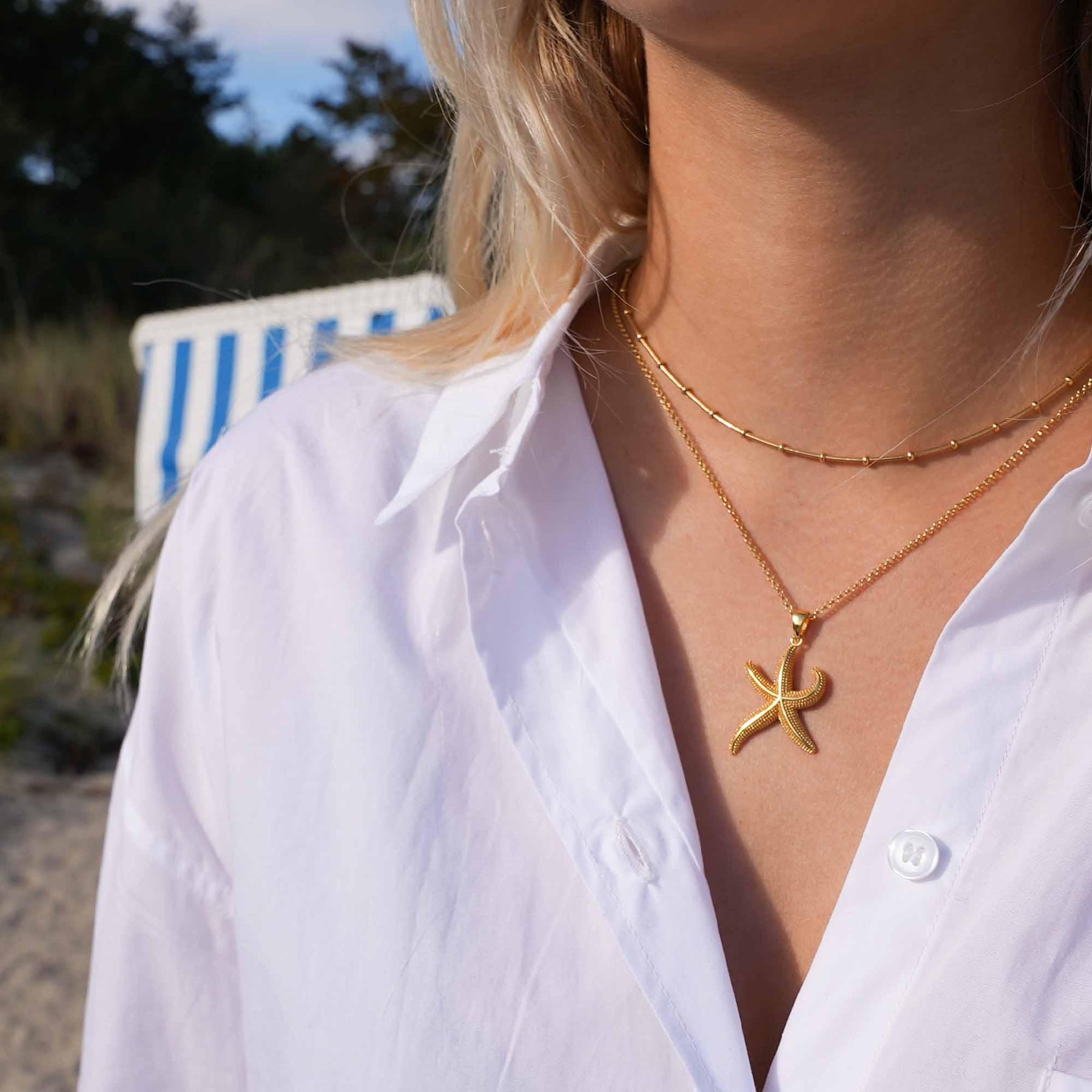 Frau am Strand mit goldener Seestern-Kette und weißer Bluse, maritimer Schmuck