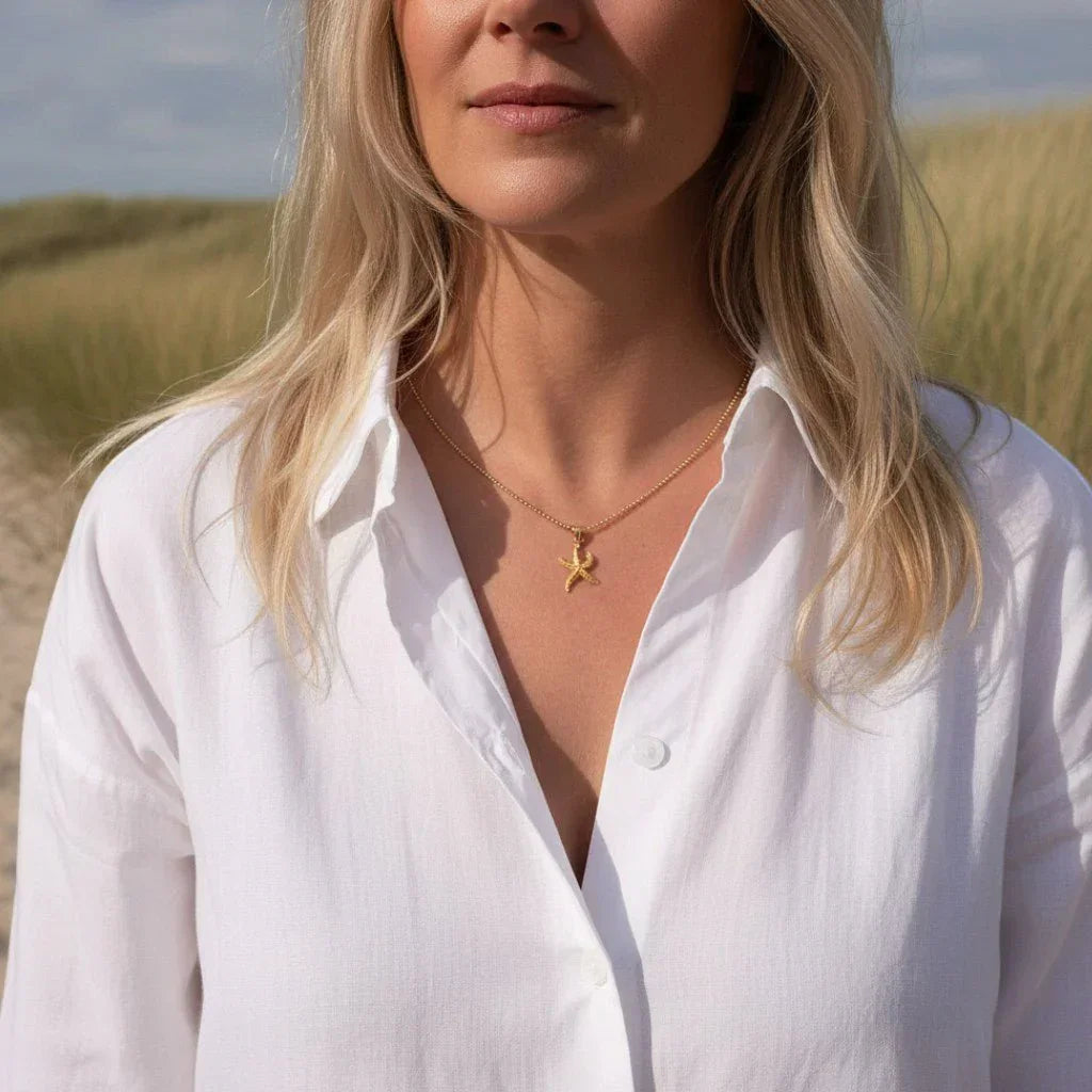 Frau mit goldener Seestern-Kette, weißer Bluse am Strand, maritimer Schmuck.